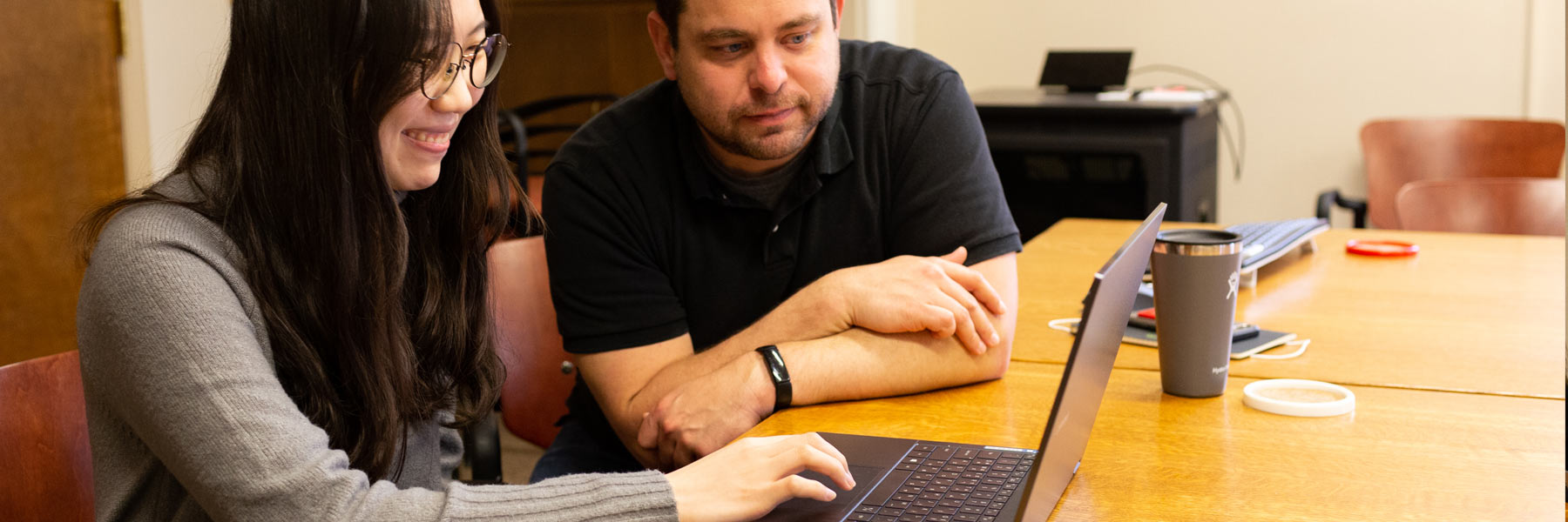 A smiling woman wearing glasses types on her laptop. Her male colleague to her right looks at the screen with her.